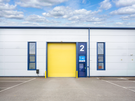 A modern industrial unit with a yellow roller shutter door labelled 2, blue-framed windows, To Let sign, and empty car park.