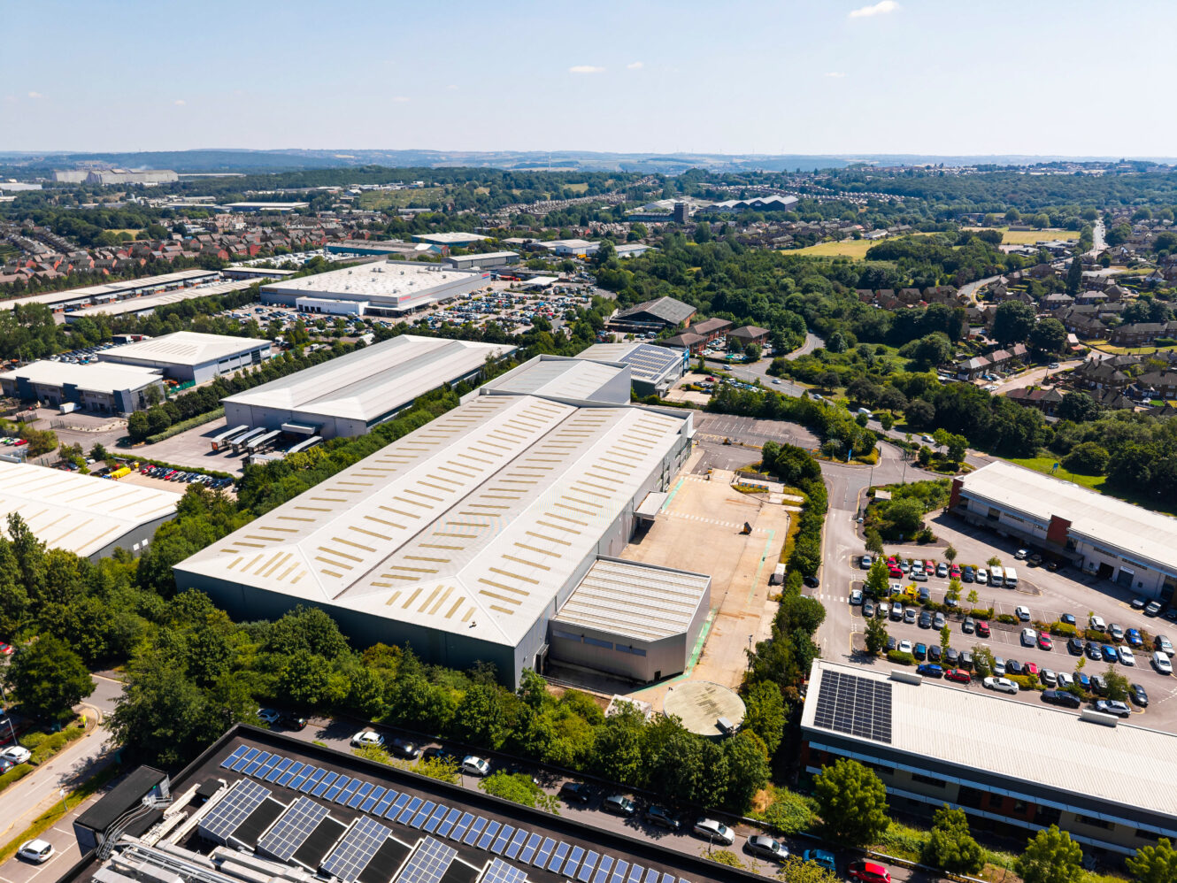 Aerial view of an industrial estate featuring large warehouses, car parks, and greenery under a clear sky.