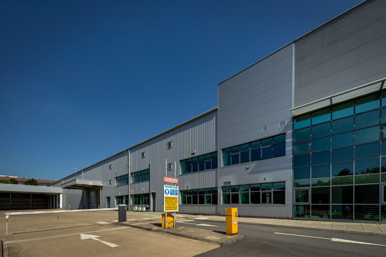 A large modern warehouse with grey metal cladding, glass windows, signs, and barriers near the entrance under a clear blue sky.