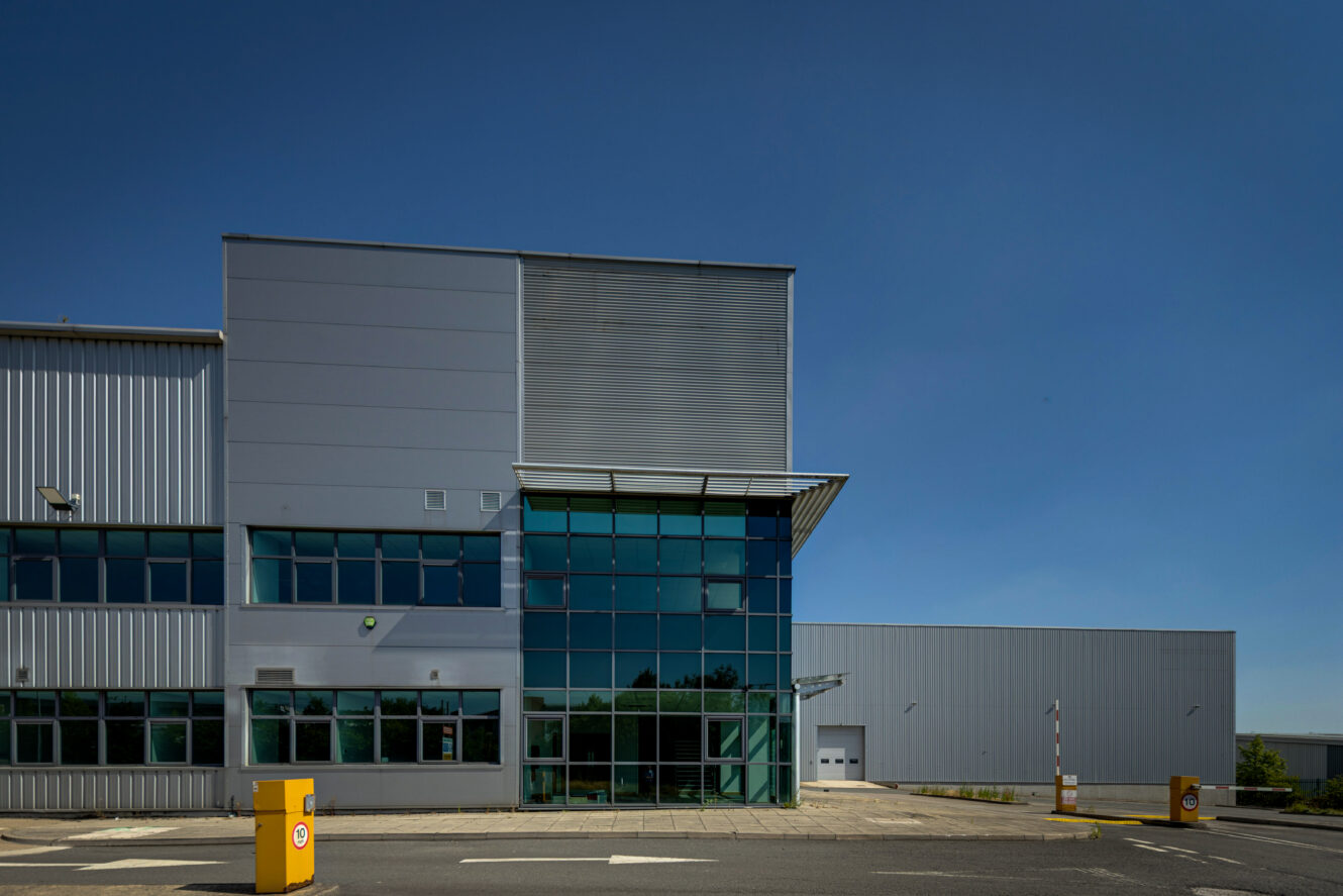 A modern industrial warehouse with large glass windows, metal cladding, and a blue sky. A yellow barrier is near the entrance.
