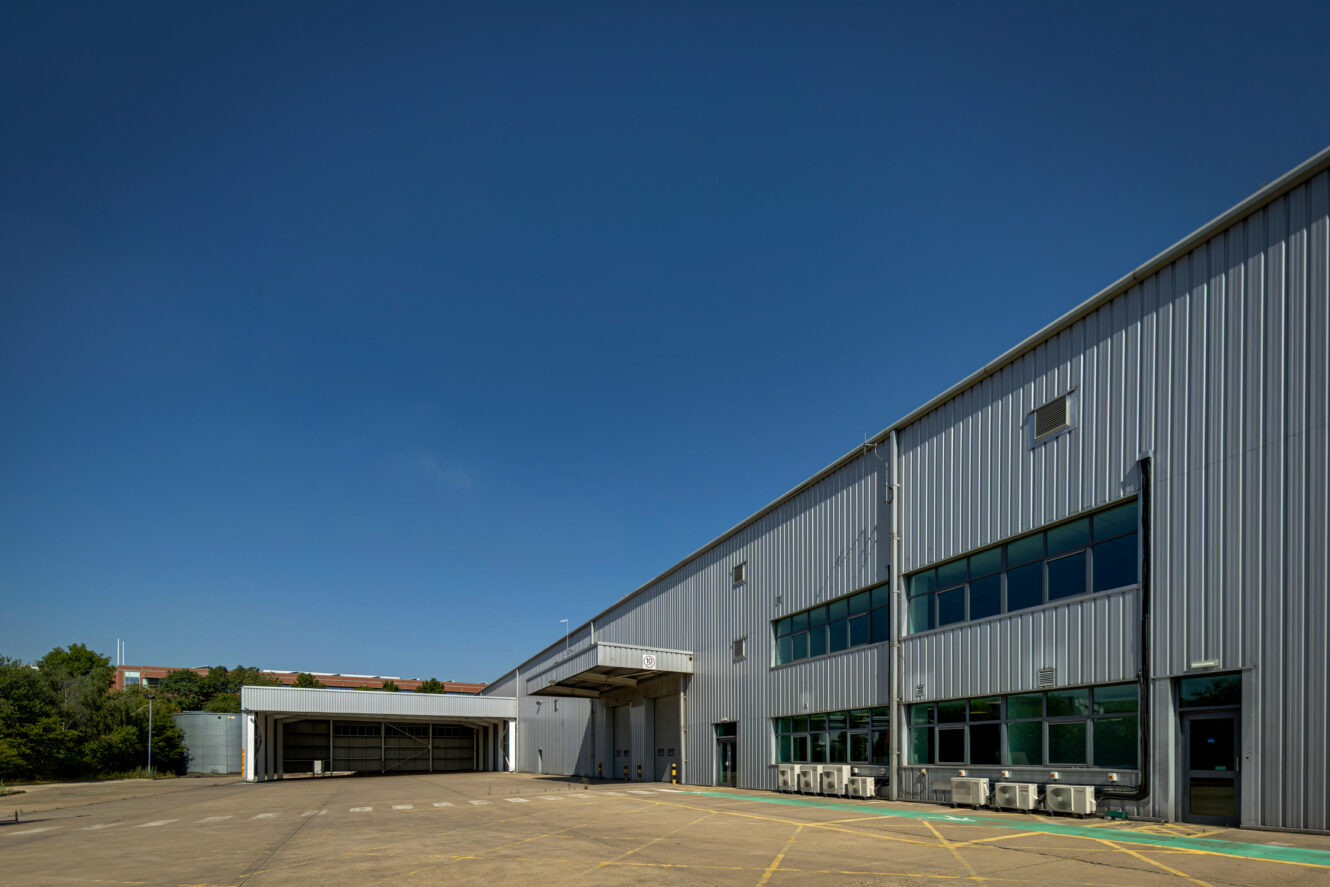 Large industrial warehouse with metal cladding and multiple windows under a clear blue sky, fronted by an empty tarmacced yard.