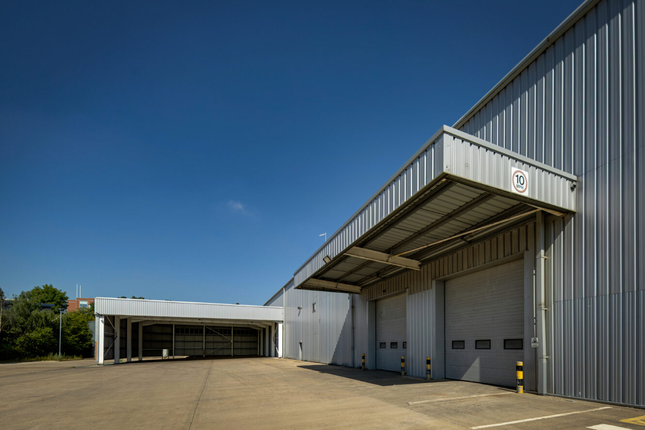A modern industrial warehouse with closed roller shutters and a canopy, beneath a clear blue sky.