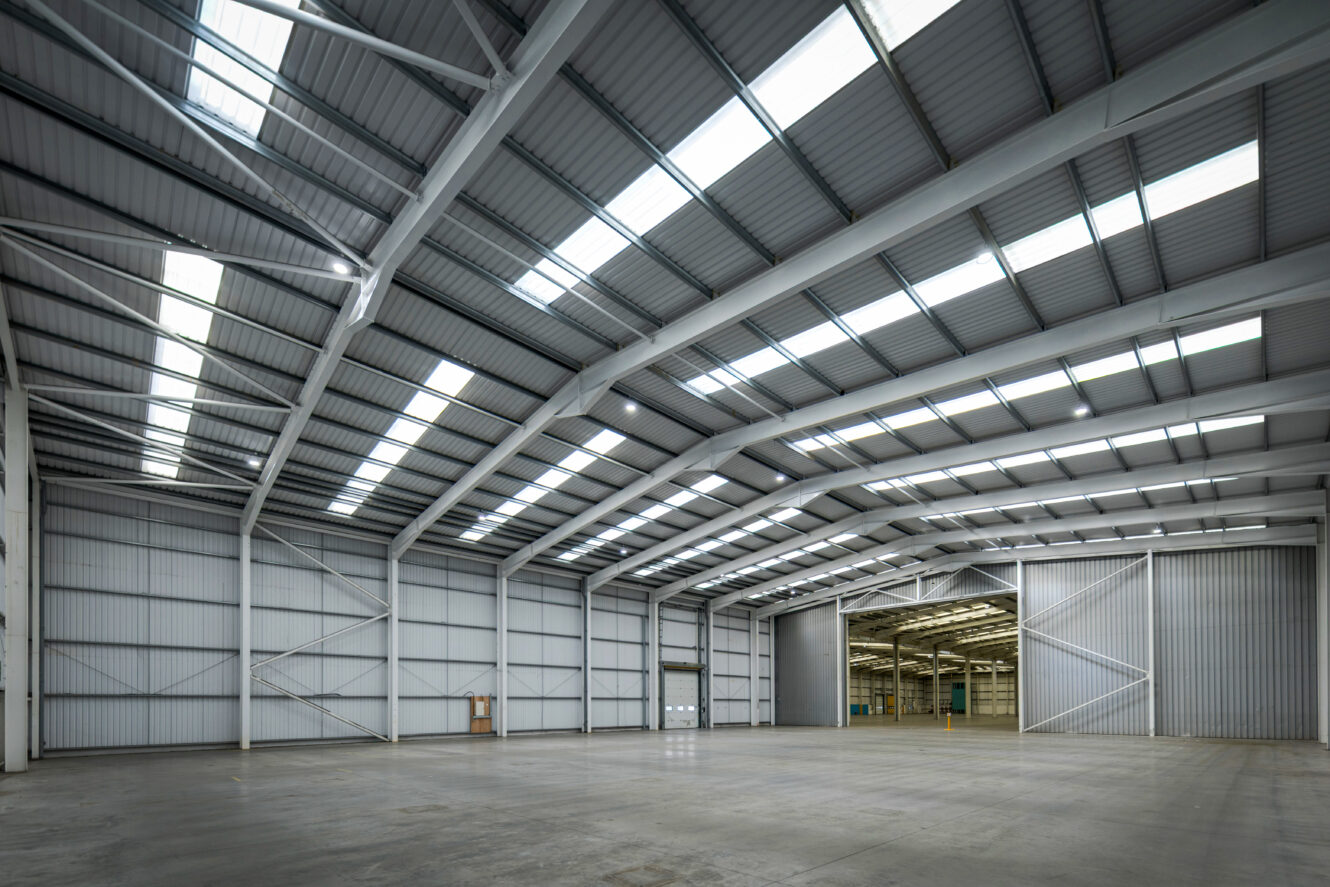 Large, empty industrial warehouse with high ceilings, skylights, concrete floors, and wide open entrance doors revealing more space.