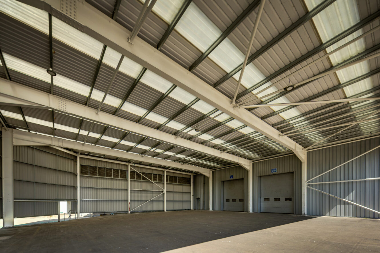 Interior of an empty industrial warehouse with high metal ceiling, large overhead doors, and concrete floor lit by natural light.