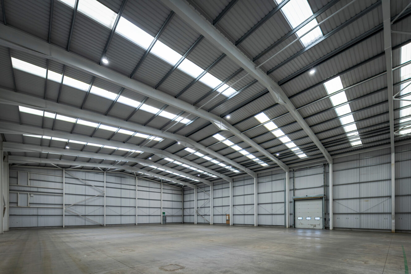 Interior of an empty industrial warehouse with high ceilings, metal walls, large skylights, and a closed roller shutter door.