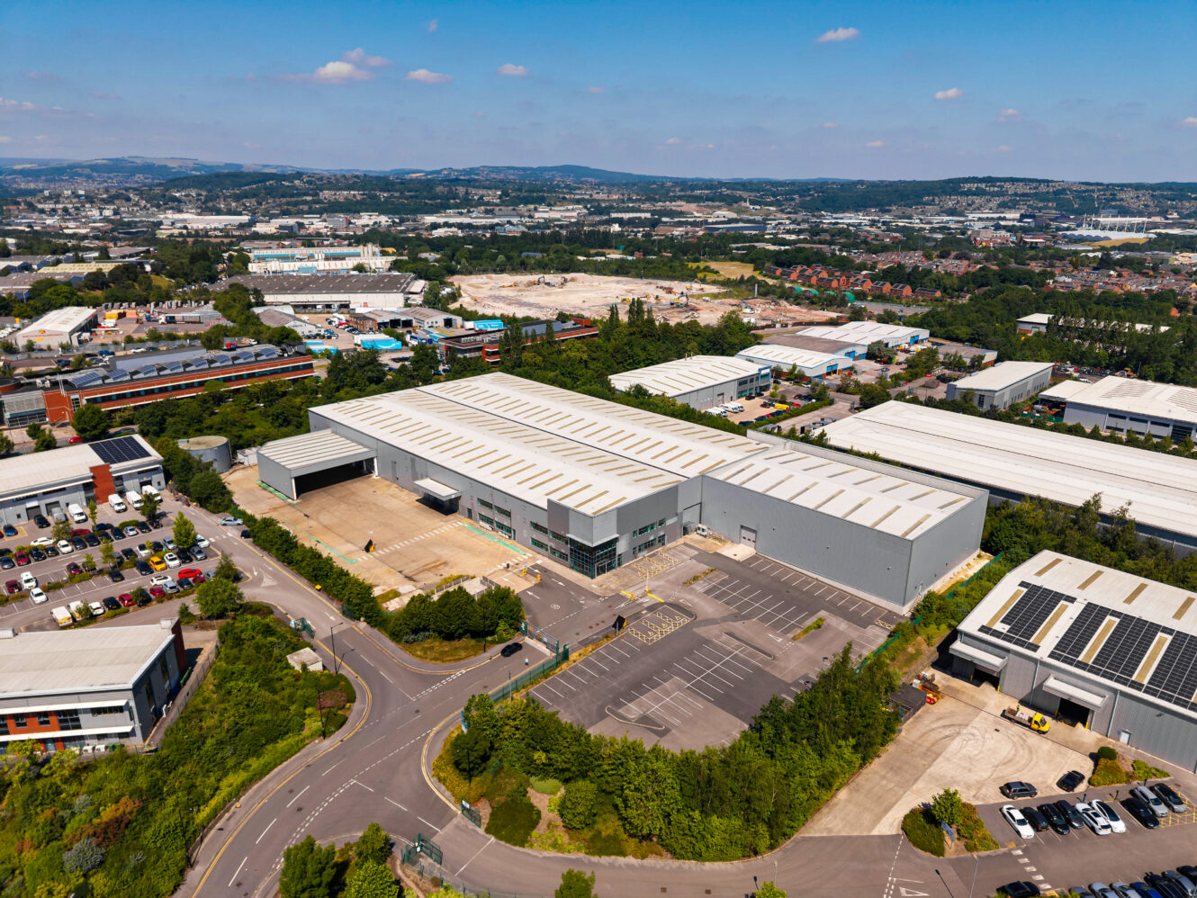Aerial view of a large industrial warehouse with loading bays, parking areas, and nearby roads in a commercial district under a clear sky.