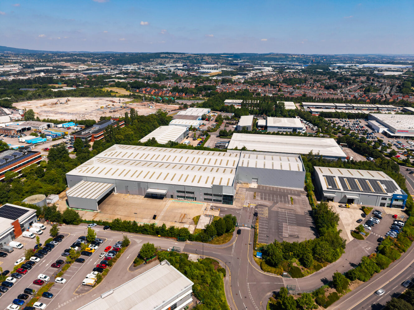 Aerial view of a large industrial warehouse complex with car parks, greenery, and nearby commercial buildings under a blue sky.