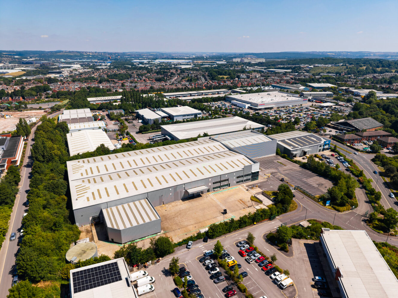 Aerial view of an industrial estate with large warehouses, car parks, and green areas nearby under a clear sky.