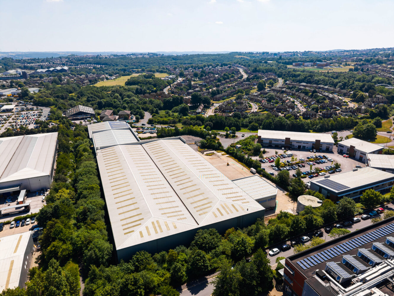 Aerial view of a large industrial warehouse among trees, with office blocks, car parks, and homes in the background.
