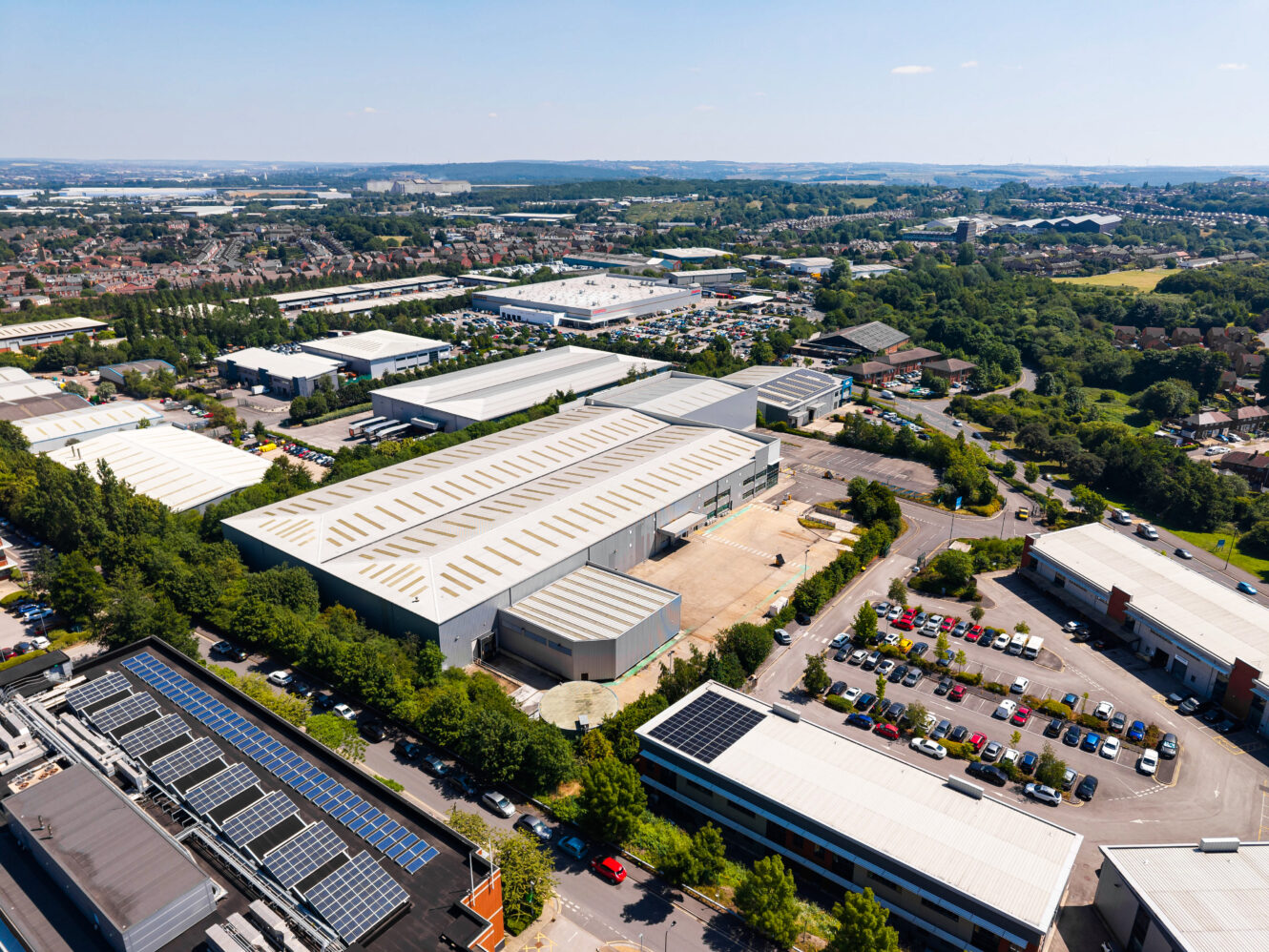 Aerial view of an industrial estate with warehouses, car parks, rooftop solar panels, and greenery under a clear sky.