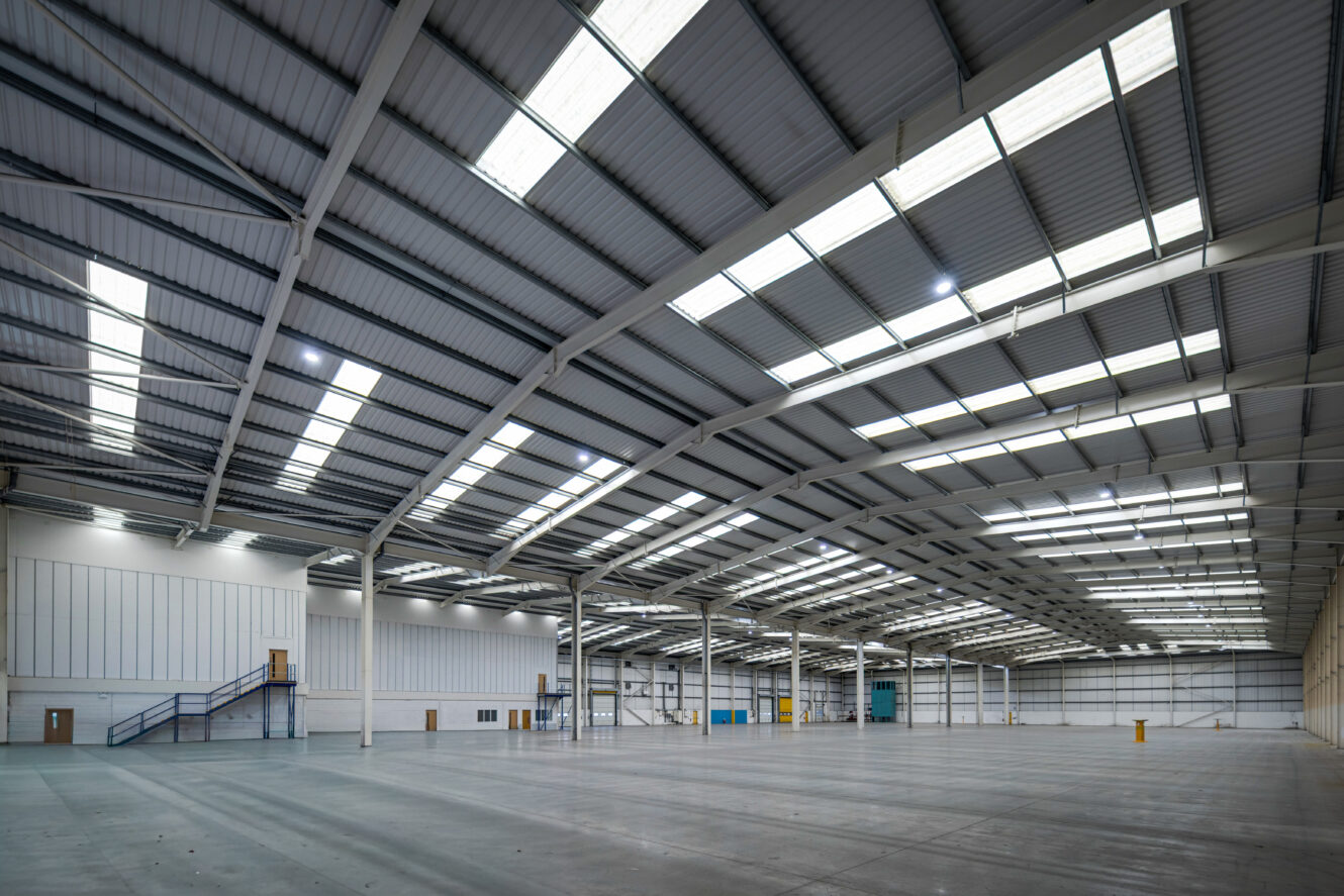 Large warehouse with high metal ceiling, skylights, concrete floor, and a staircase to a mezzanine on the left; no people present.