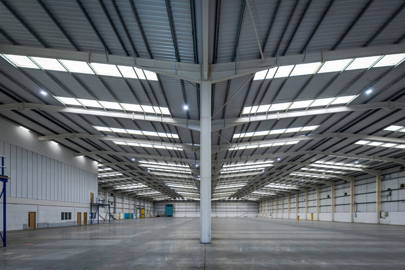 Interior of a large, empty warehouse with high ceilings, metal beams, skylights, and concrete floor. Industrial doors line the walls.