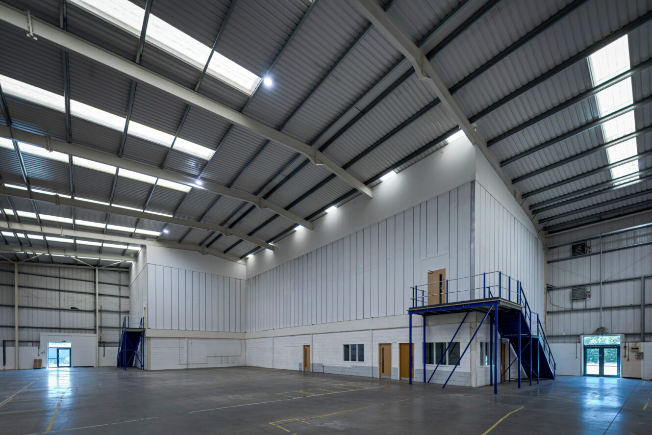 Spacious, empty warehouse interior with high ceilings, metal beams, and a raised office on the right with stairs and blue railings.