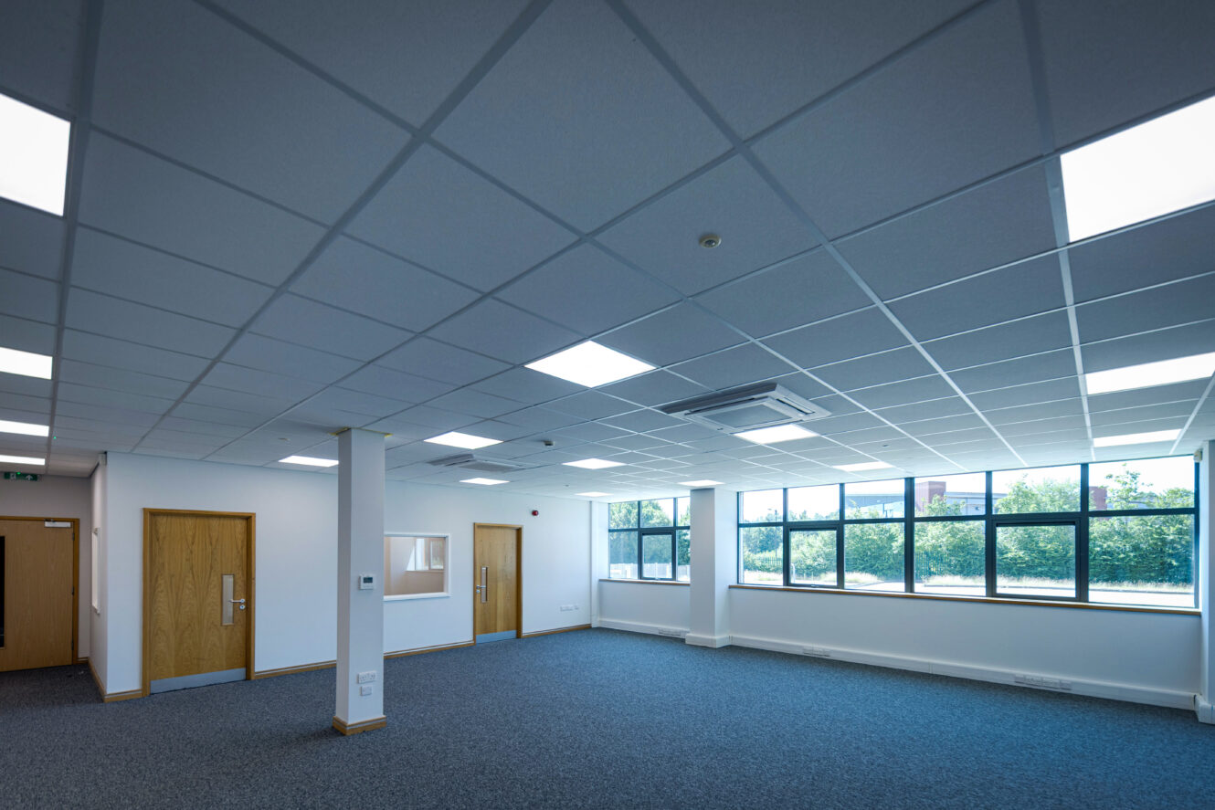 Empty office space featuring carpeted floors, white walls, wooden doors, large windows with natural light, and recessed ceiling lights.