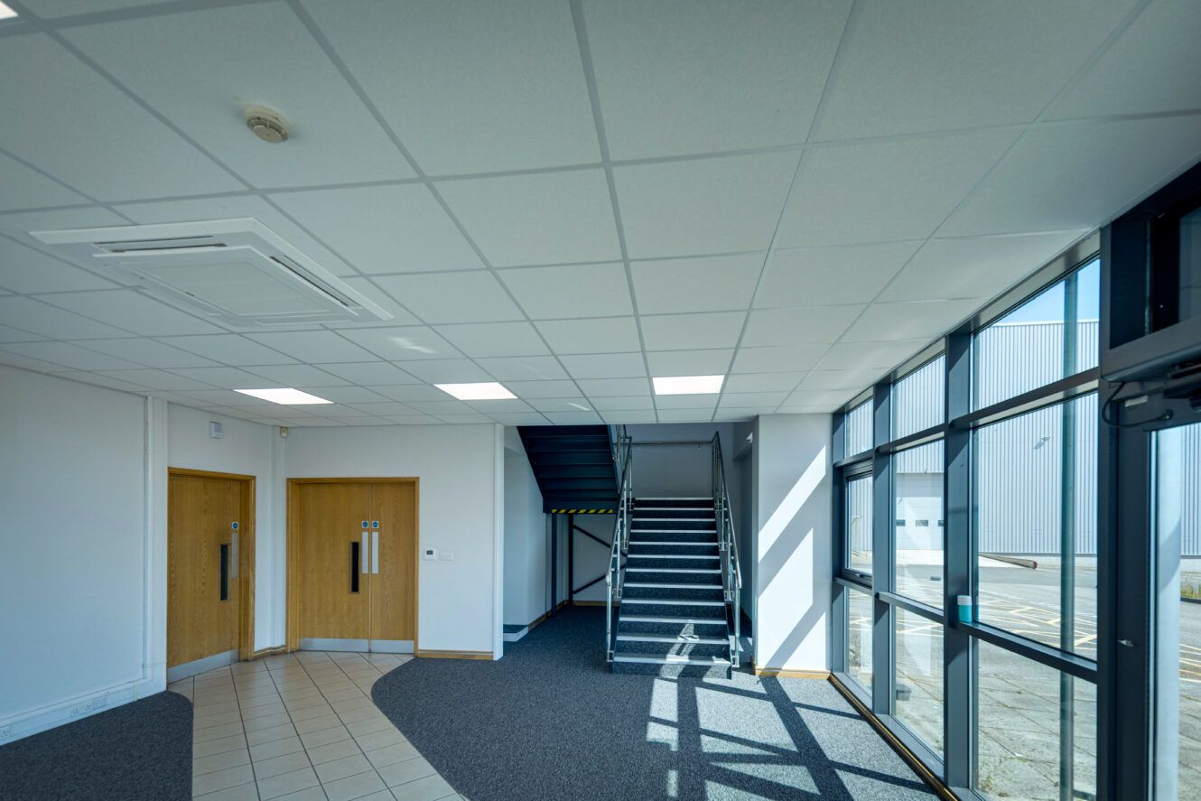 A modern office lobby with tiled and carpeted floors, large right-side windows, two wooden doors, and a central staircase upwards.