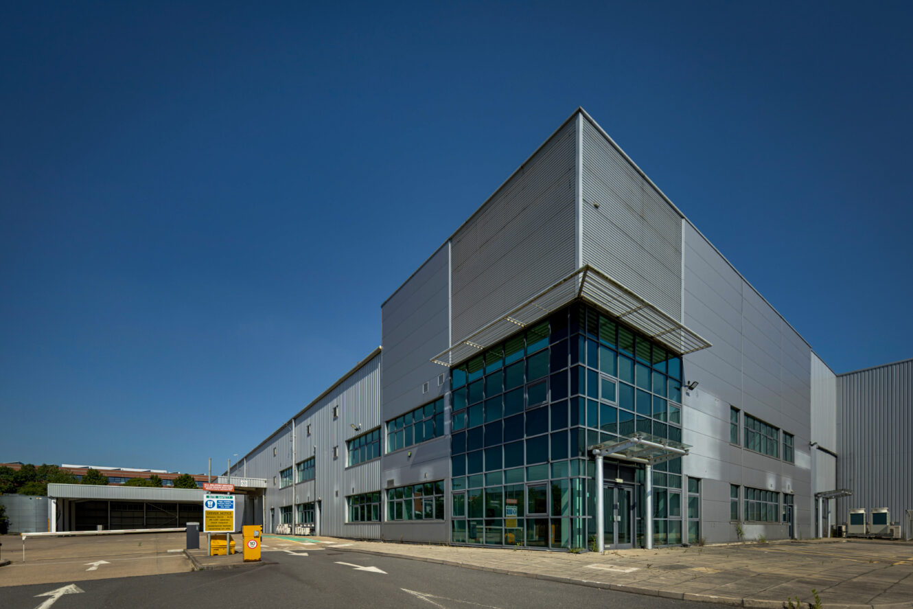 A large, modern industrial warehouse with glass entrance walls, clear blue sky above, and a small car park at the front.