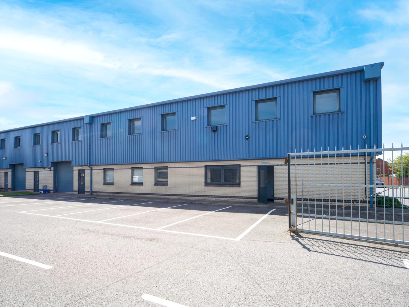 A modern industrial building with blue metal cladding and beige lower walls, behind an open gate and an empty car park under a clear sky.