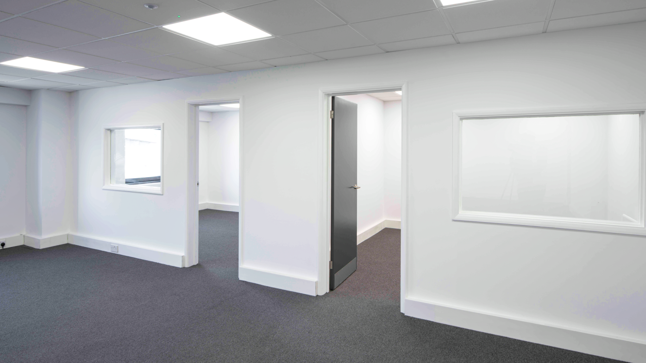 Empty office space featuring white walls, grey carpet, ceiling lights, two open doorways, a closed door, and internal windows.