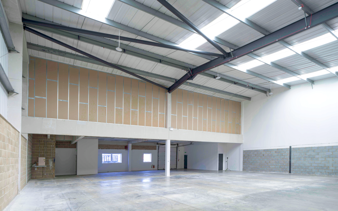 Large empty warehouse with high ceilings, exposed beams, concrete floors, unfinished interior walls, and windows and doors at the back and sides.