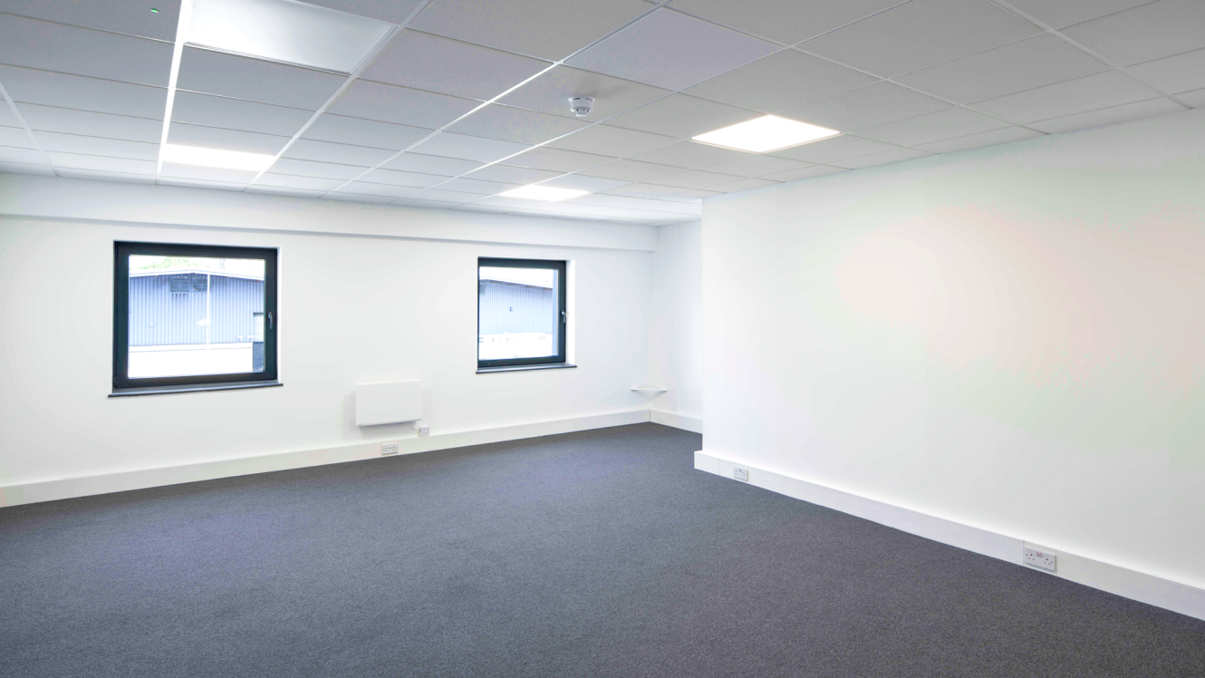 Empty office room with white walls, two windows, grey carpet flooring, and overhead ceiling lights.