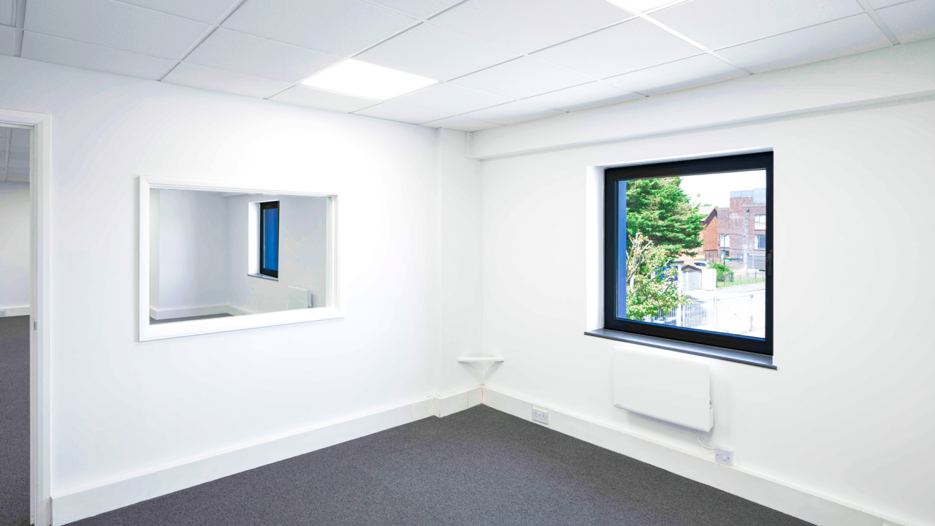 An empty office with white walls, a window showing buildings and trees, a large wall mirror, and grey carpeted floors.