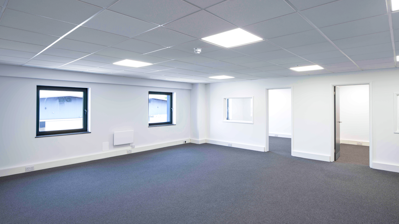 Empty office room with two windows, a small internal window, and two open doors. White walls, grey carpet, fluorescent lights overhead.
