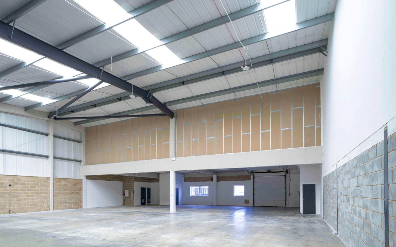 Interior of an empty industrial warehouse with high ceilings, exposed beams, large windows, and concrete floors.