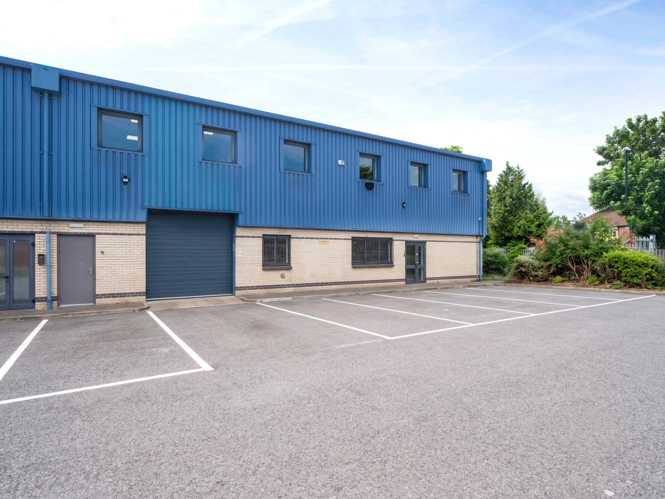 A blue and beige industrial building with multiple windows and a large garage door, facing a newly marked empty car park.