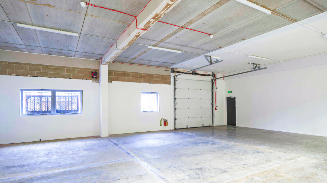 Empty industrial warehouse with concrete floors, white walls, high ceiling, two windows, a large garage door, and a fire exit.