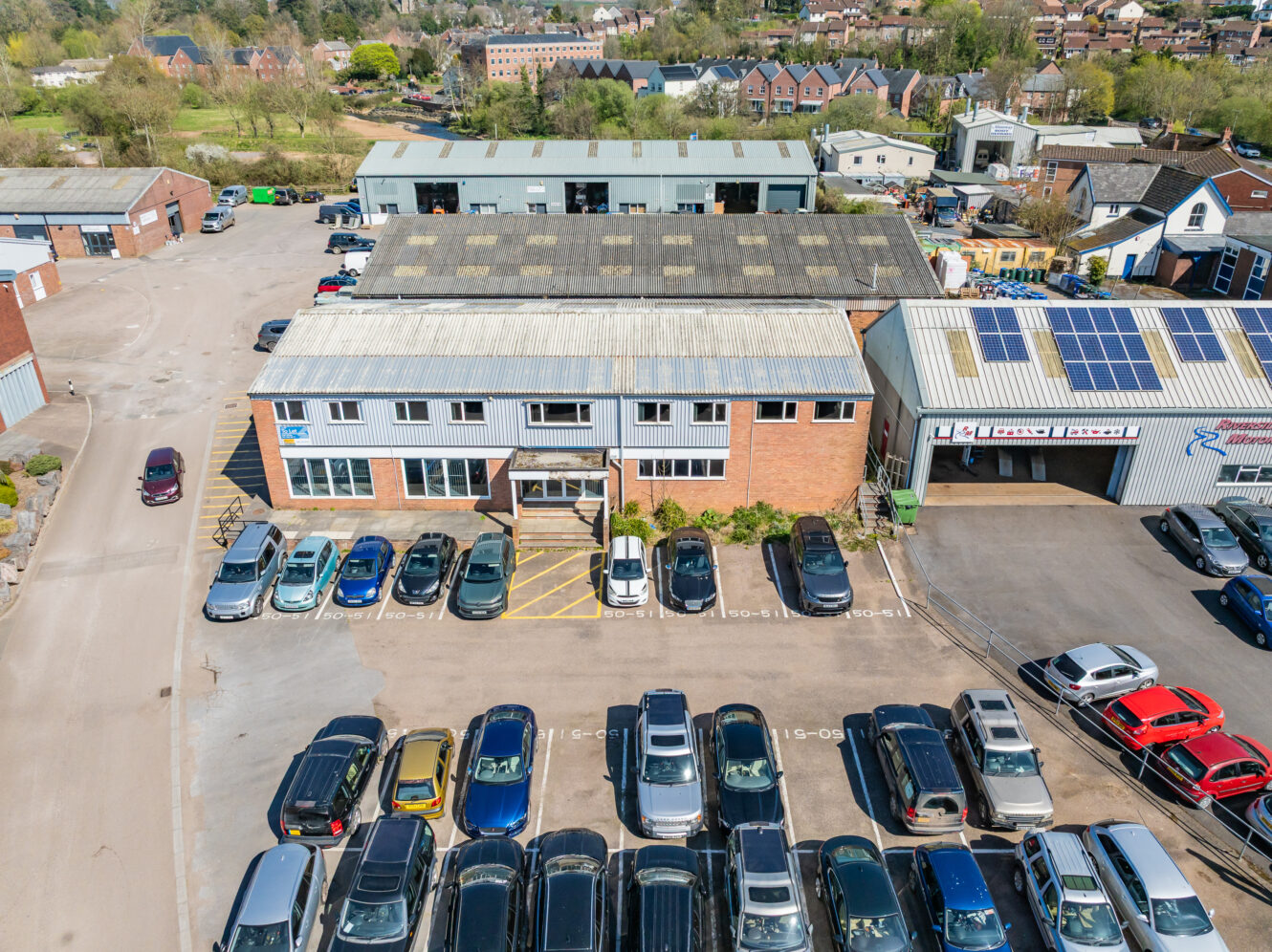Aerial view of a commercial building with a busy car park; rooftop solar panels visible on another nearby building.