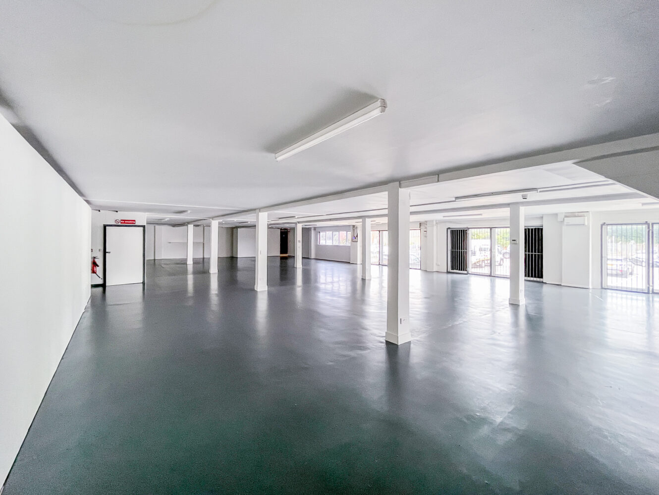 Spacious empty room with white walls, support columns, polished dark floor, fluorescent lights above, and large windows on one side.