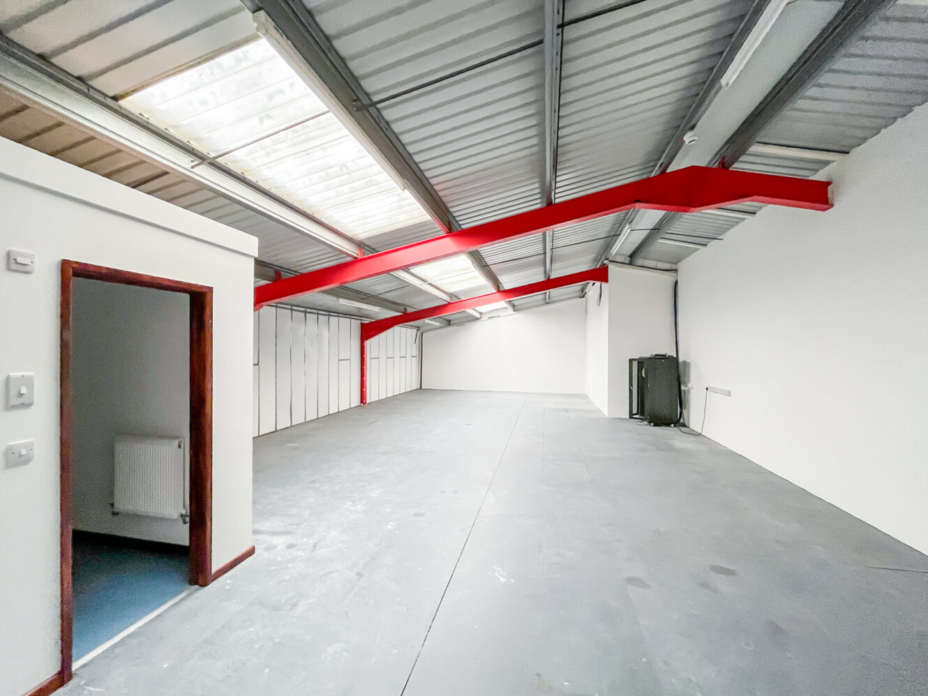 Empty industrial warehouse with grey concrete floor, white walls, red ceiling beams, skylights, and a small room with open door.