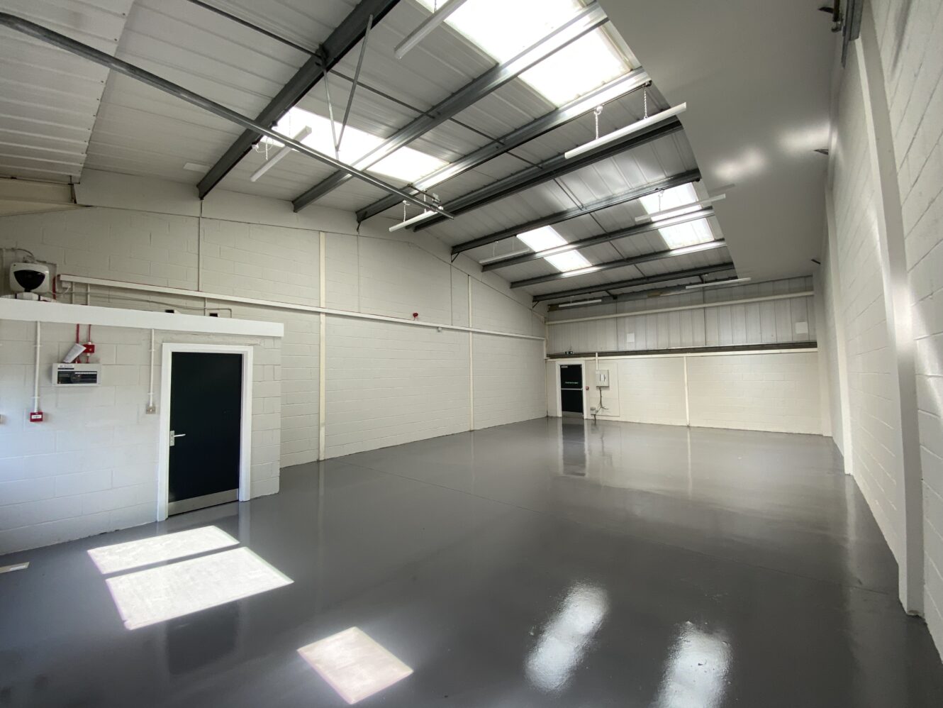 Empty warehouse with white walls, grey epoxy floors, high ceiling, and skylights. Two black doors and electrical panels are visible.