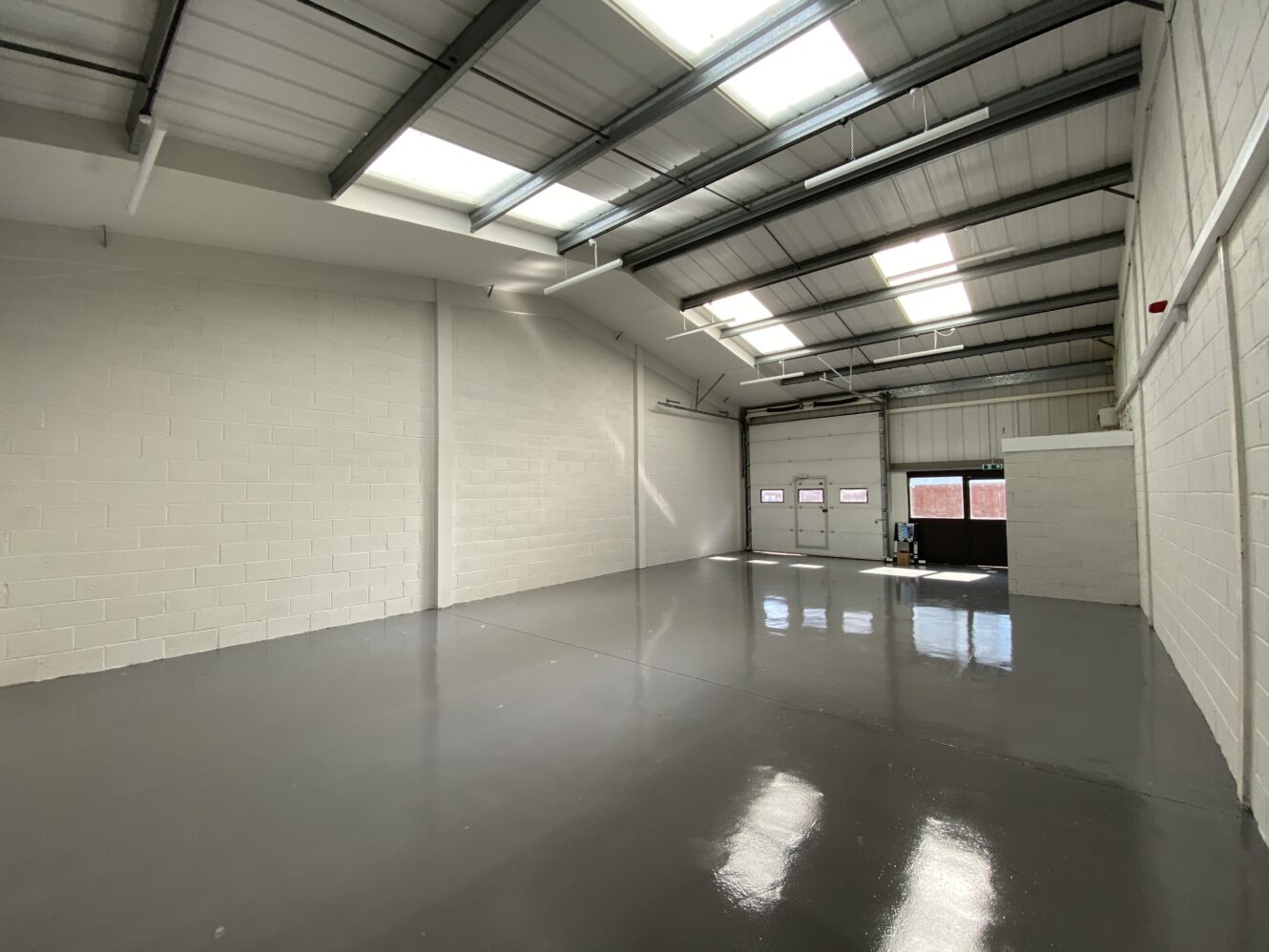 An empty industrial warehouse with white walls, polished grey floor, and a metal ceiling with skylights. Large doors and windows ahead.