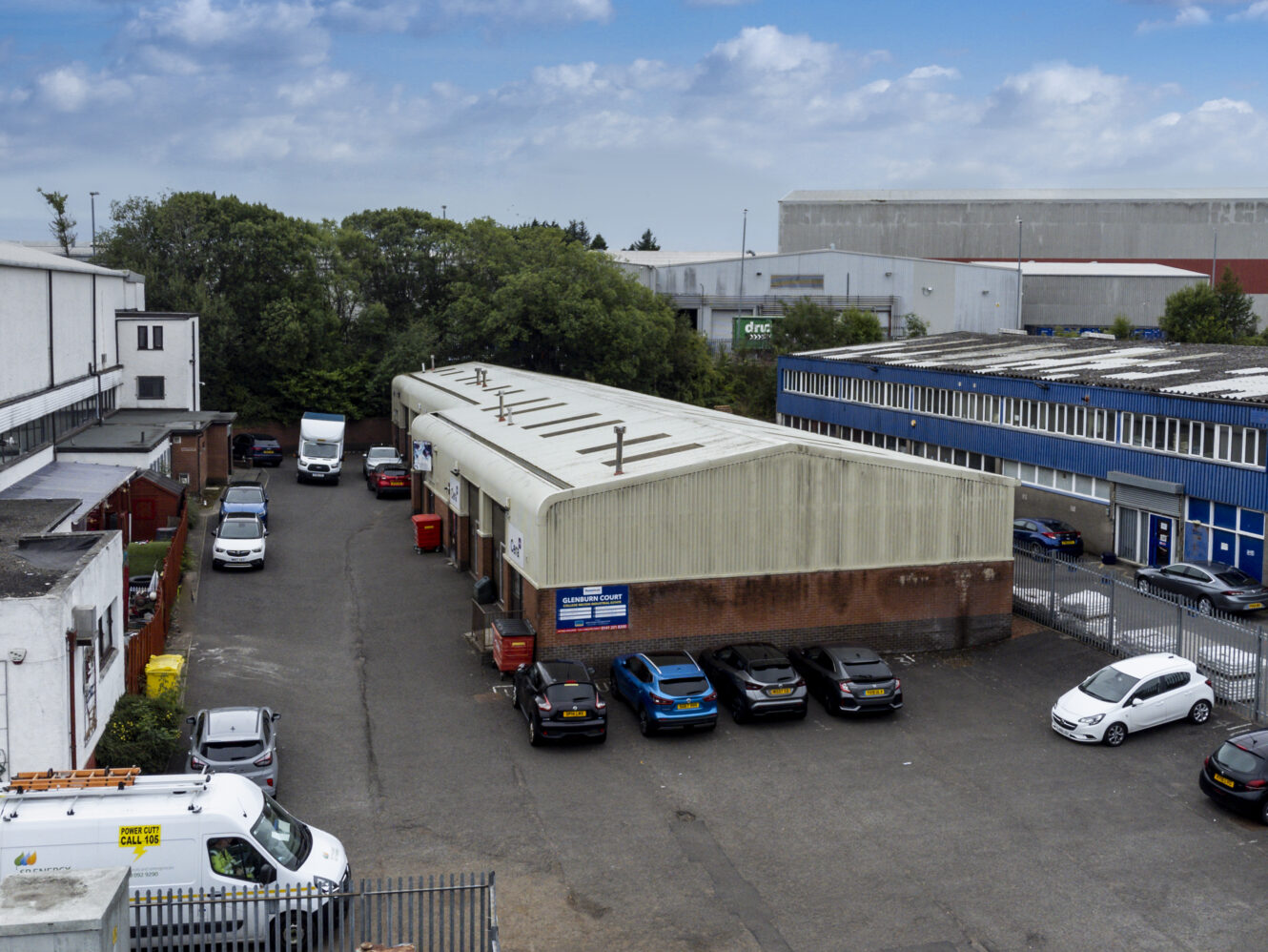 Industrial yard with parked cars and vans near warehouse buildings, trees, and fencing beneath a partly cloudy sky.