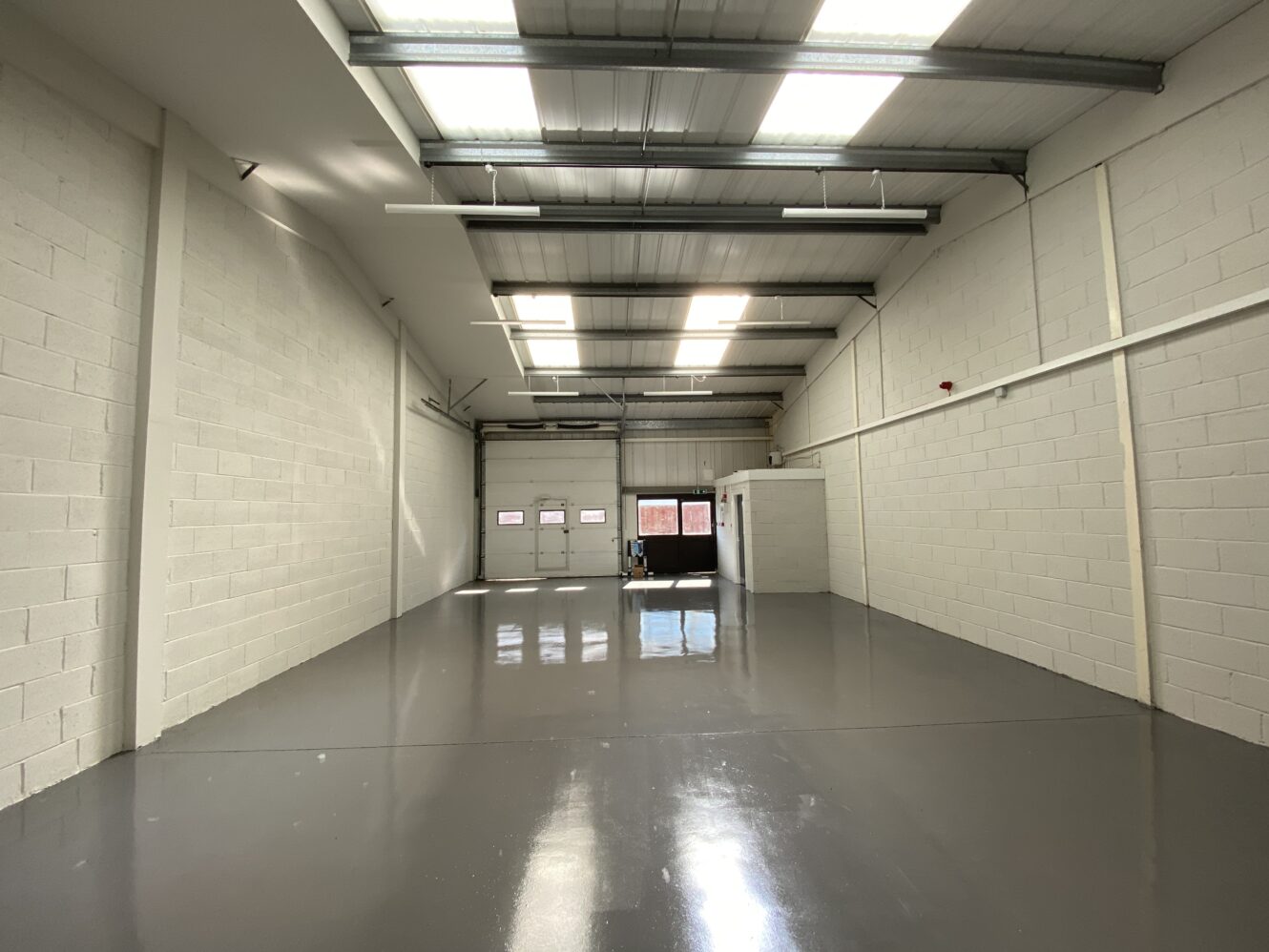 Empty industrial warehouse with white brick walls, glossy grey floor, high ceiling, fluorescent lights, and a large roller shutter door.