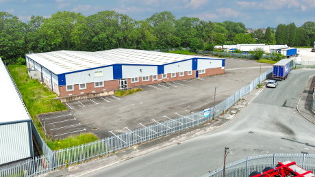 A large, empty warehouse with blue and white exterior, adjacent car park, metal fence, in an industrial area with trees behind.