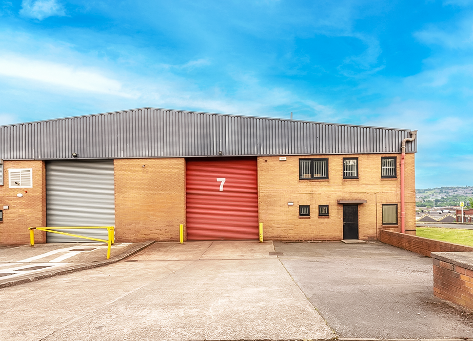 A large industrial warehouse with a red roller shutter labelled 7, a grey roller shutter, and a sloped drive under a blue sky.