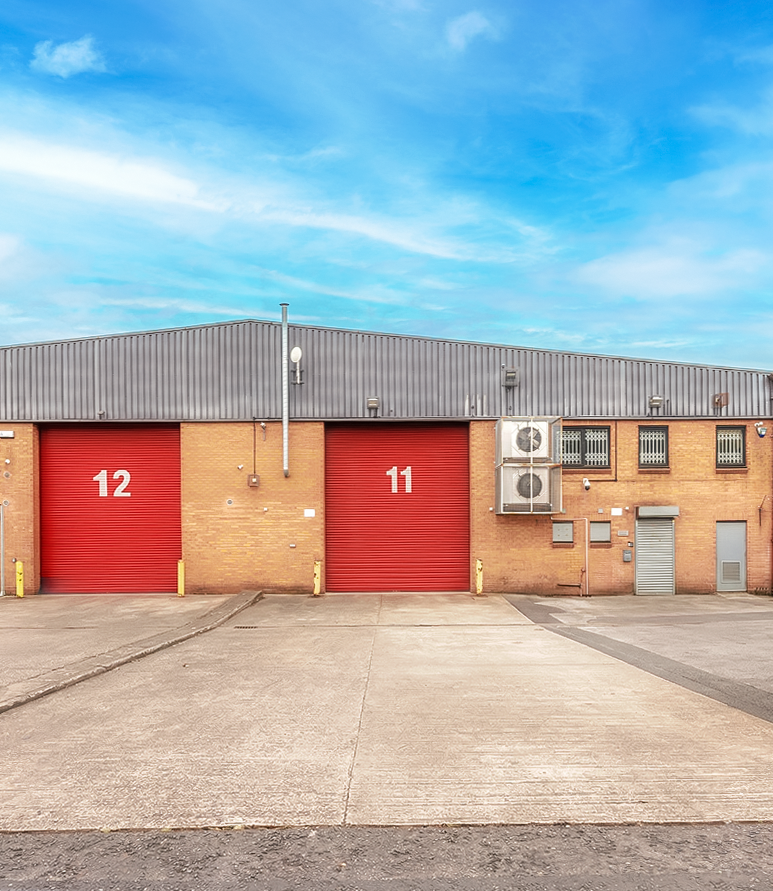 A brick industrial warehouse with two large red doors labelled 11 and 12, air conditioning units, and a concrete driveway below a blue sky.