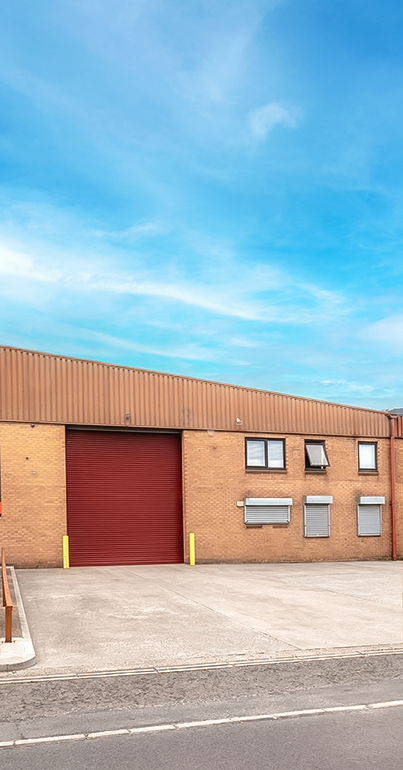 A brown brick industrial building with a large red roller shutter, several windows, and a clear blue sky overhead.