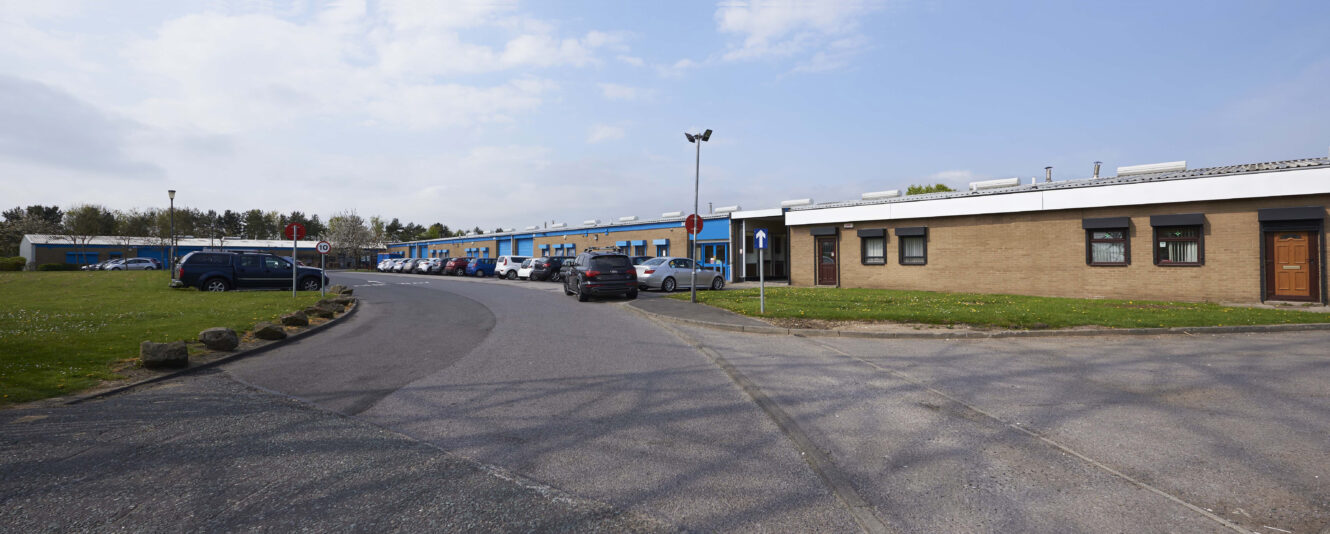 A curved drive leads to a low industrial building with blue and brown sections, parked cars, and grass under a partly cloudy sky.