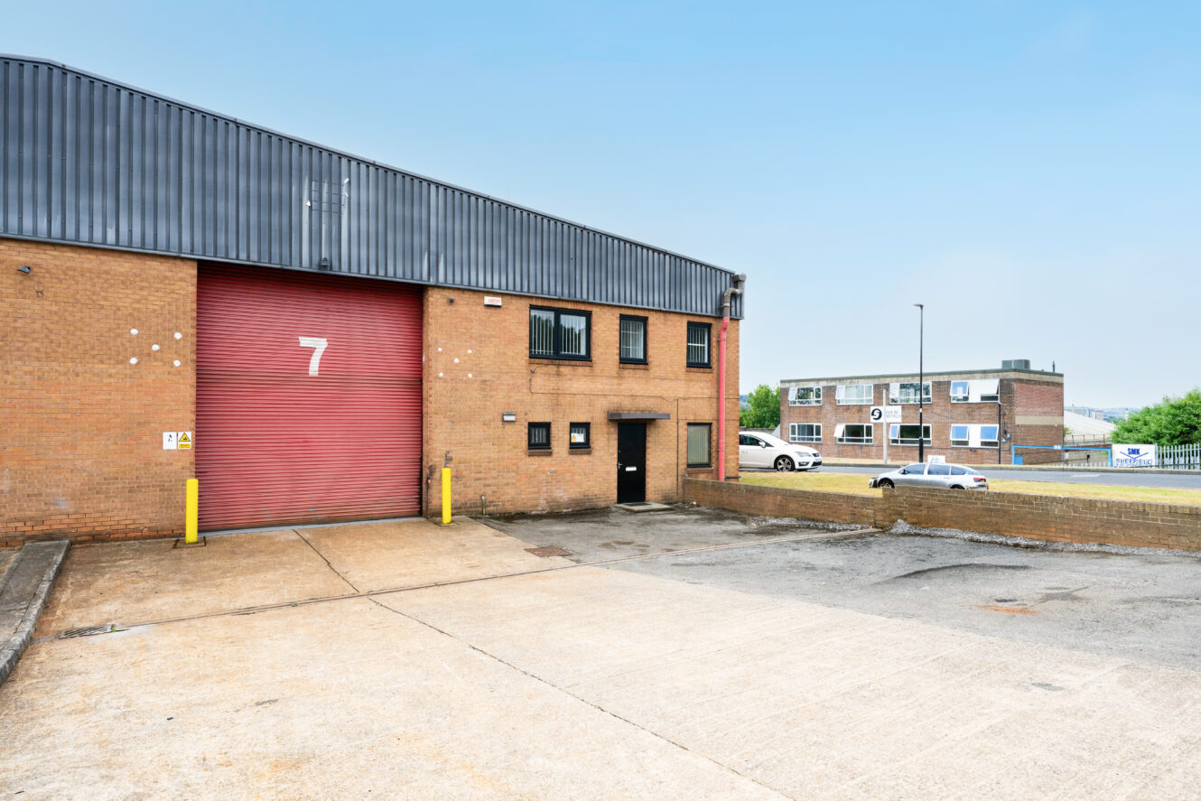 A brown brick industrial building with a red roller shutter marked “7” and an empty concrete yard; office and cars in the background.
