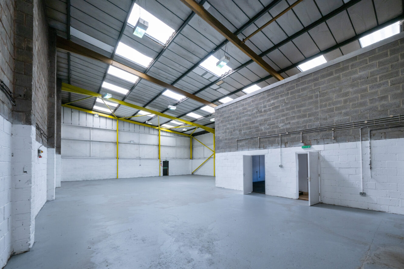 Interior of an empty warehouse with high ceilings, exposed beams, concrete floor, and white brick walls; open doorways lead to smaller rooms.