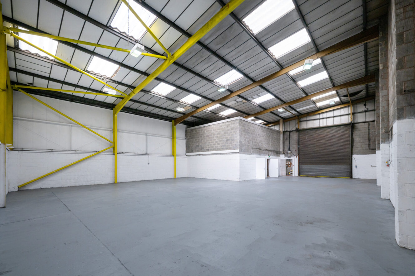 Empty industrial warehouse with high ceilings, yellow support beams, white brick walls, concrete floor, and a large roller shutter door.