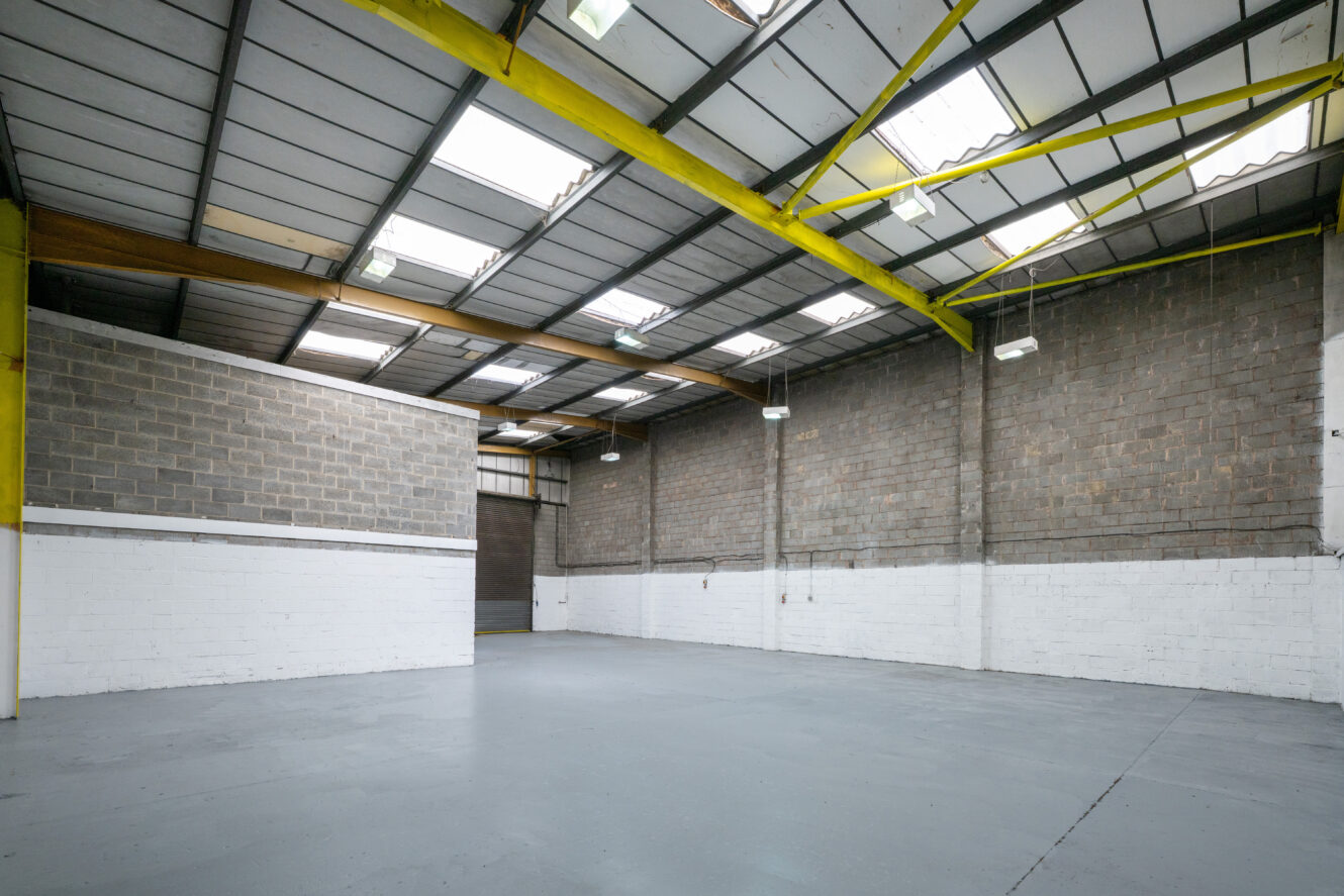 Empty industrial warehouse with high ceilings, concrete floor, brick walls, and skylights letting in natural light.