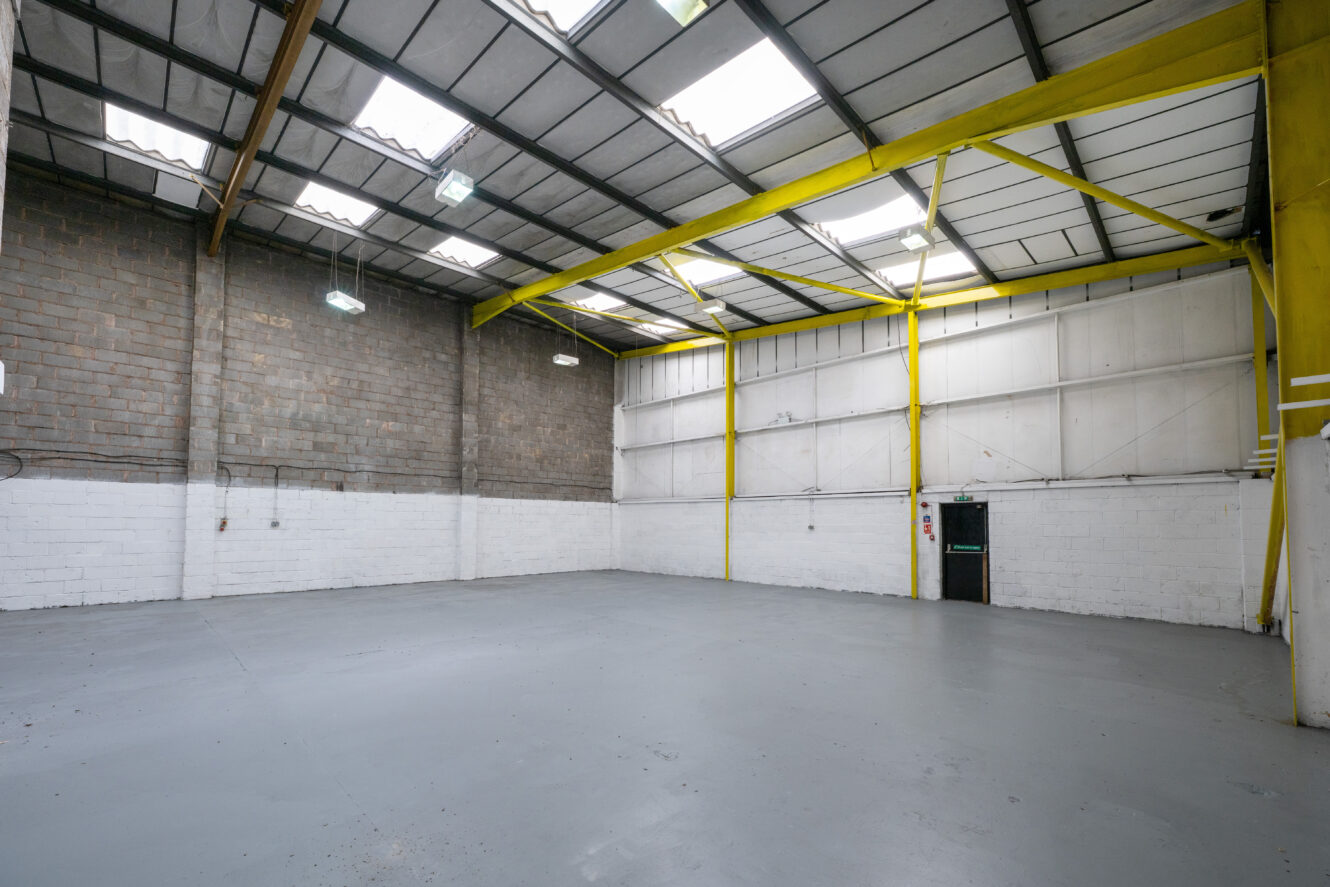 Empty industrial warehouse with high ceilings, concrete floor, brick and white walls, yellow beams, fluorescent lights, black door.