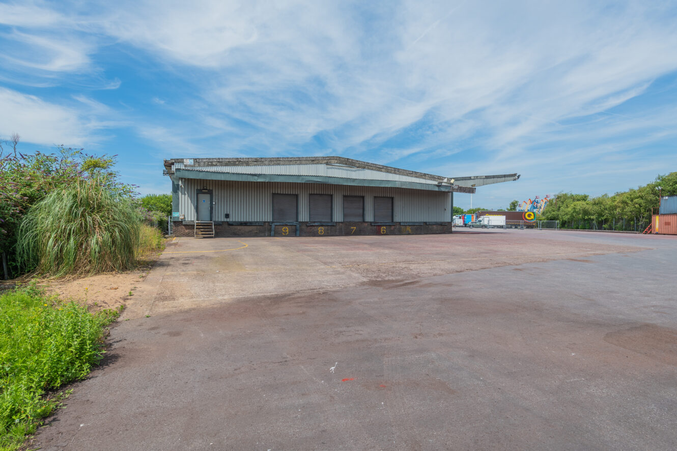 A large, empty loading bay with numbered bays sits beside an open tarmacked area and greenery beneath a blue sky.