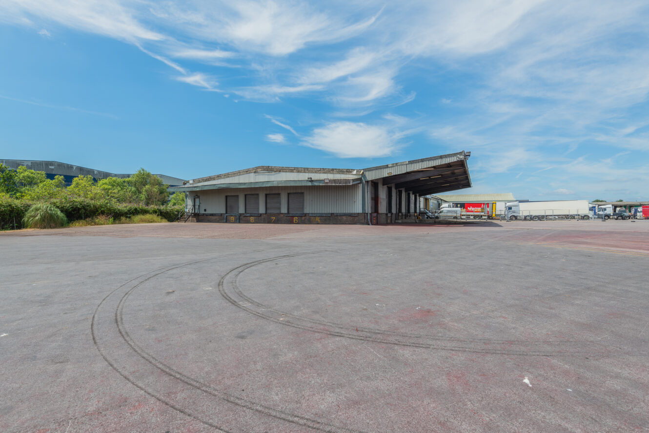 A large, empty loading bay with tyre marks on concrete and lorries in the background under a blue sky with scattered clouds.