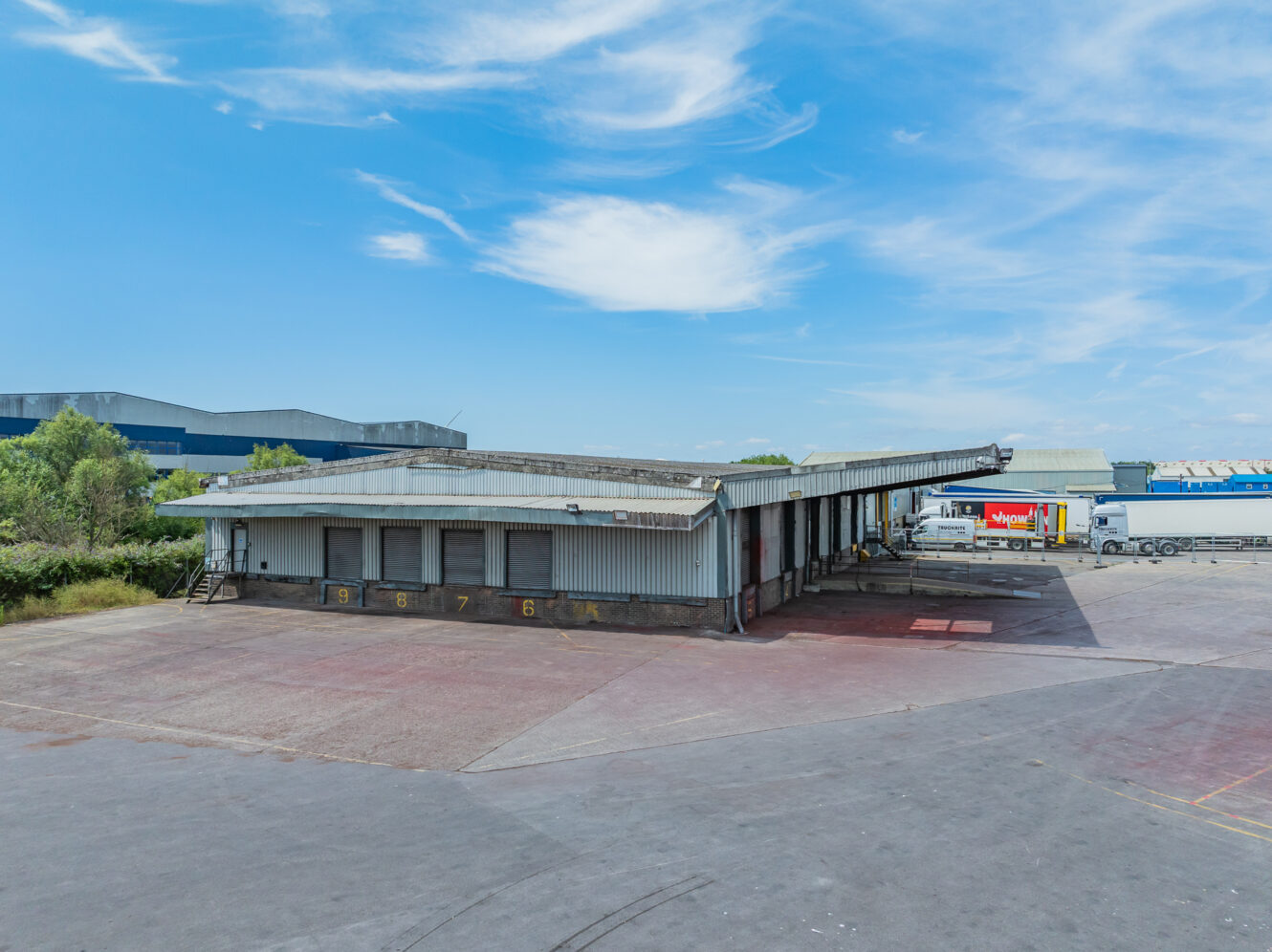 A large, empty industrial loading bay with numbered bays, next to a tarmacked car park and warehouses under a partly cloudy sky.