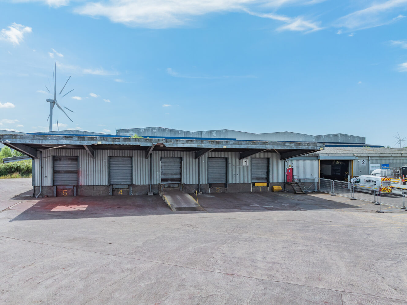 A row of five loading dock bays at an industrial warehouse on a clear day, with a wind turbine and metal fencing in the background.